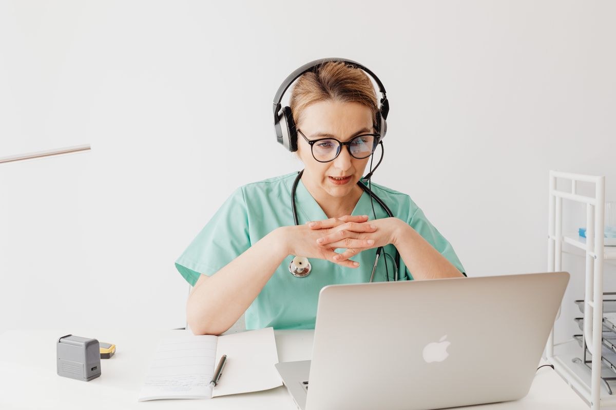 A healthcare professional in mint-green scrubs on a video call with a patient on a laptop — virtual homeopathy consultation